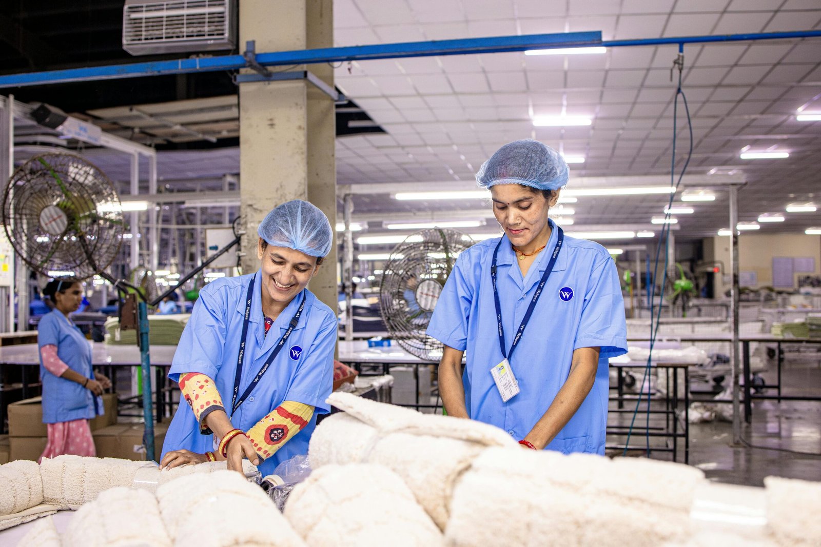 Smiling Indian factory workers in a textile factory wearing blue uniforms and hairnets, working diligently.